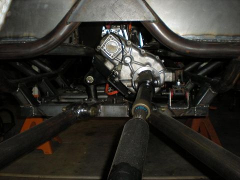 Suzuki Samurai - Underside view looking forward at a tube-chassis buggy on jack stands, showing the aluminum transfer case mounted between the frame rails w…