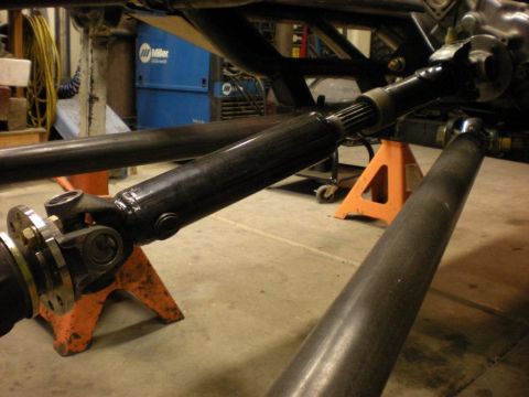Suzuki Samurai - A glossy black driveshaft with universal joints and a flanged yoke propped on orange jack stands in a fabrication shop, with raw tube stock…