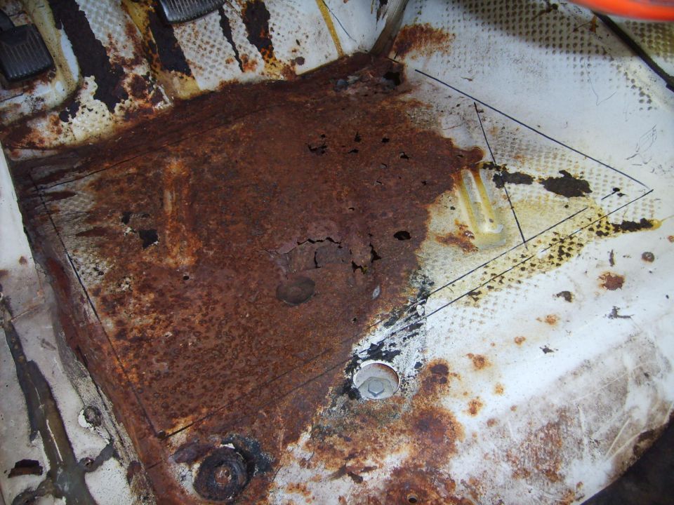 Heavily rusted car floor pan with holes, pedals visible, and repair panel outlines drawn on white floor.