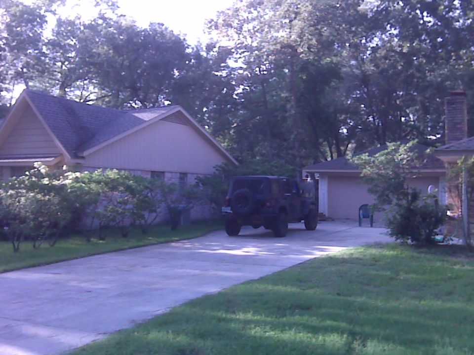 Dark Jeep Wrangler with rear-mounted spare parked on a driveway by a house and garage