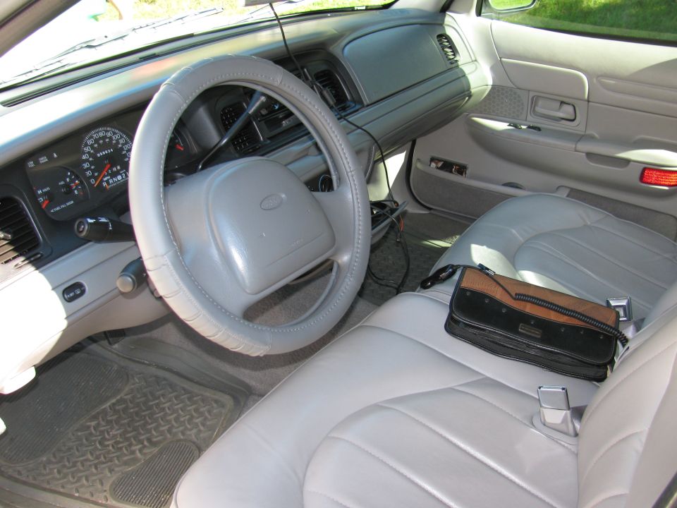 Gray interior of a Ford sedan showing steering wheel with cover, gauges, front bench seat and a case on the passenger seat