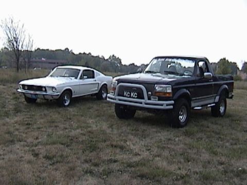 1995 Ford Bronco - A black Ford pickup truck and a white Ford Mustang are parked side-by-side on a grassy field.