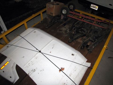 1984 Ford Bronco - A trailer carries automotive parts, including a white hood and suspension components, with a truck in the background.