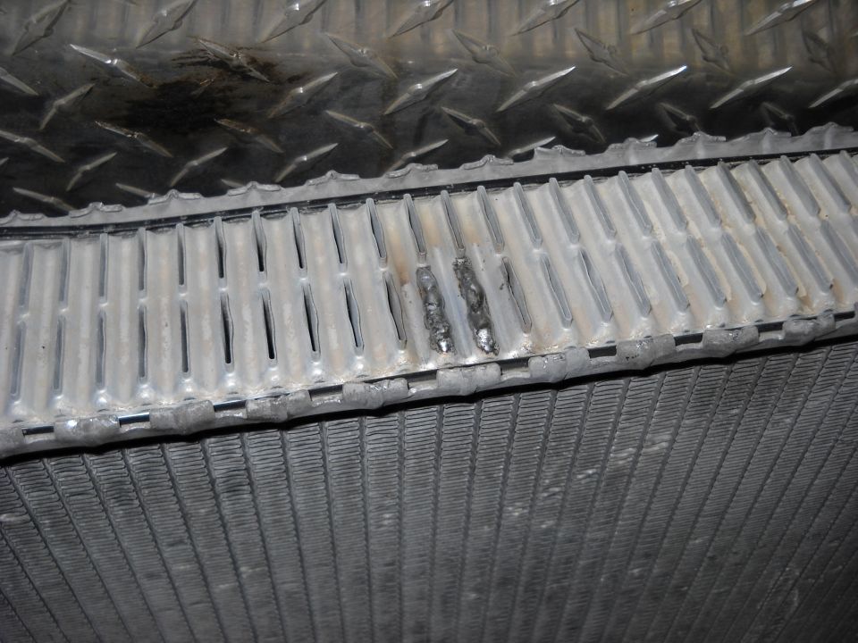Close-up of a damaged radiator with two visible weld repairs on the top fins. Diamond plate metal is above