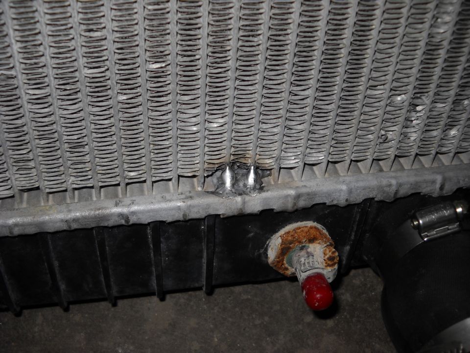 Radiator with bent fins and a dented metal tank, showing a red drain plug and black hoses