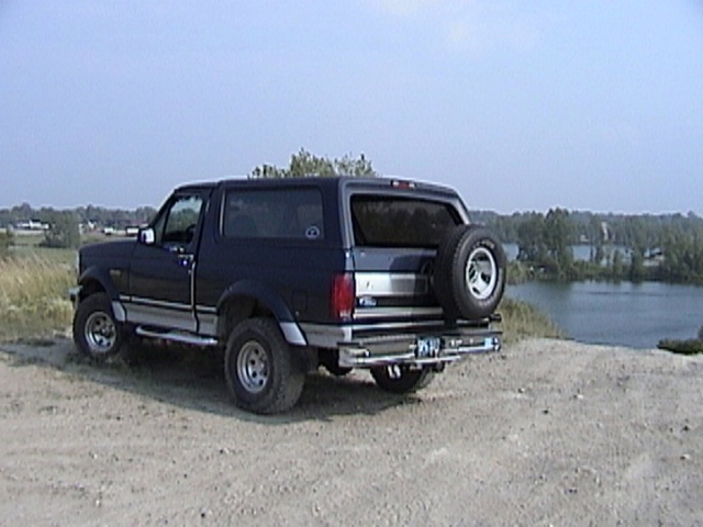 Dark blue Ford Bronco parked on dirt near a lake, rear-mounted spare tire visible
