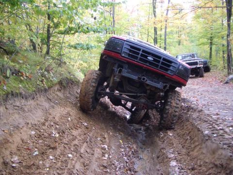 1994 Ford F-250 - A red Ford truck drives through a muddy rut on a wooded trail, with other trucks visible in the background.
