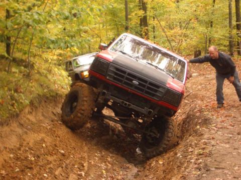 1994 Ford F-250 - A red and black Ford truck is seen off-roading with a man assisting. Another vehicle is visible behind it.