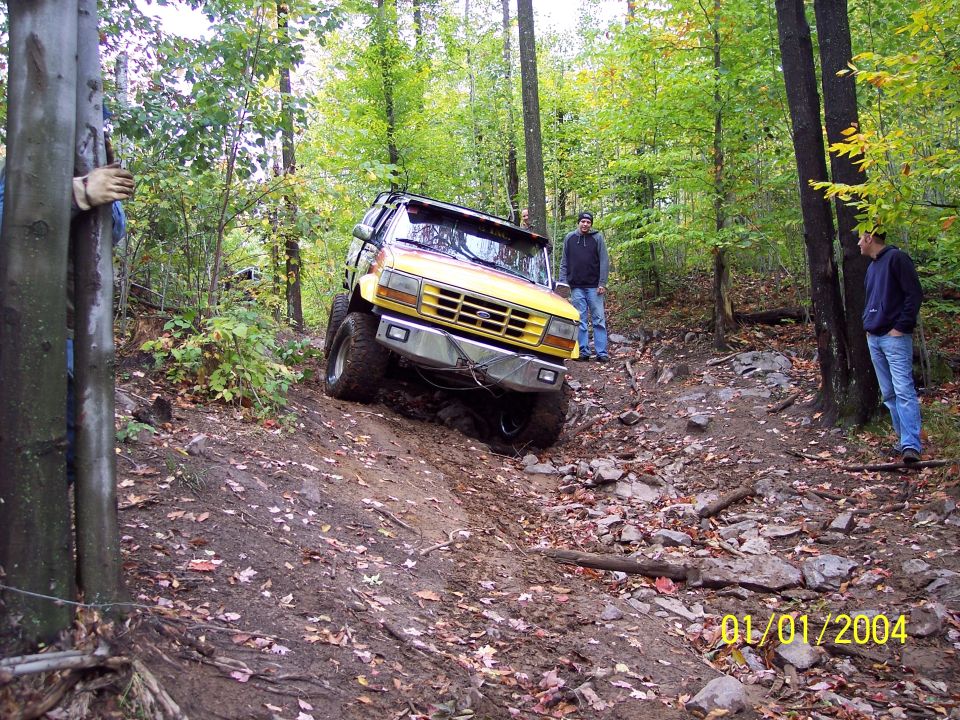 Yellow off-road truck on a muddy, rocky trail. Front bumper is chrome. Two men stand nearby