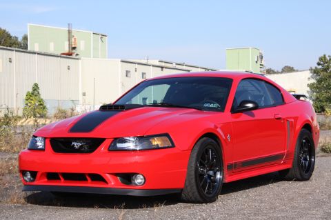 2004 Ford Mustang - A red 2004 Ford Mustang coupe is parked outside industrial buildings under a clear sky.