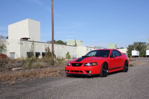 2004 Ford Mustang - A red 2004 Ford Mustang coupe is parked outside industrial buildings under a clear blue sky.