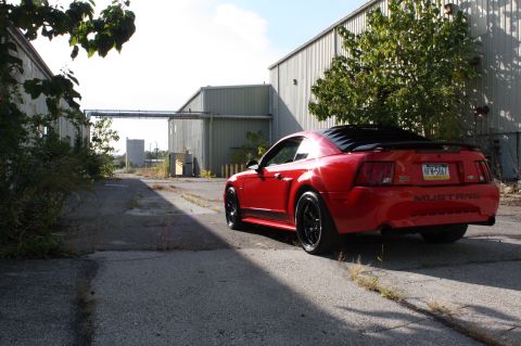 2004 Ford Mustang - A red 2004 Ford Mustang is parked in an alley between industrial buildings.