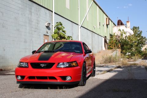 2004 Ford Mustang - A red 2004 Ford Mustang with black racing stripes is parked in front of an industrial building.