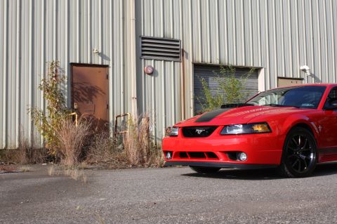 2004 Ford Mustang - A red 2004 Ford Mustang is parked in front of a warehouse-style building with overgrown weeds.