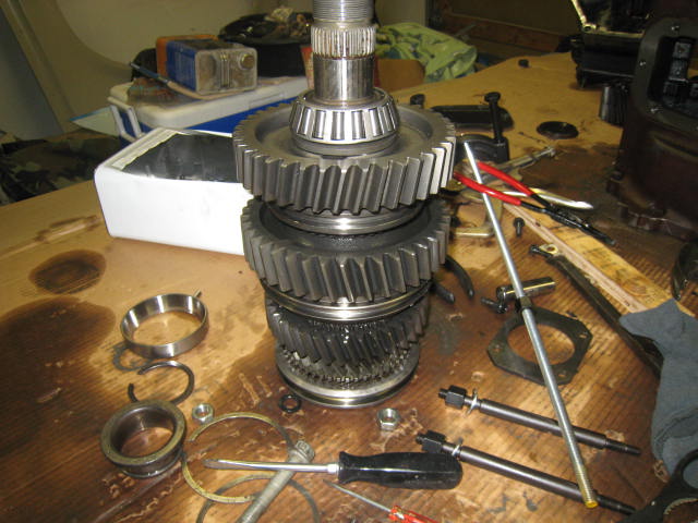 Transmission mainshaft with gears stacked upright on a workbench surrounded by bearings, shift rods, and hand tools