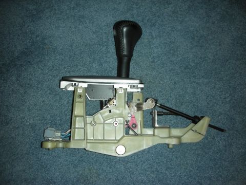 1998 Ford Crown Victoria - A detached gear shifter assembly, a black leather-topped shift knob and intricate mechanical linkages, is displayed against a blue carpeted…