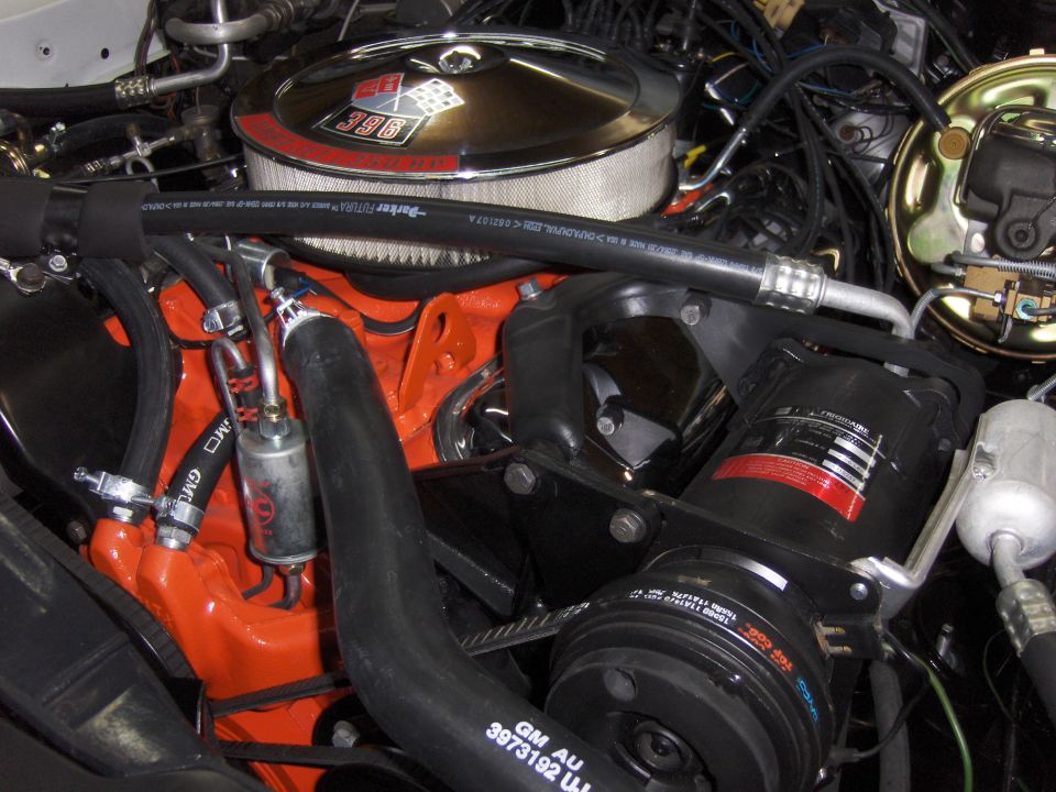 Close-up of a V8 engine with a chrome air cleaner showing a 396 badge. Orange block, black hoses, and AC compressor visible