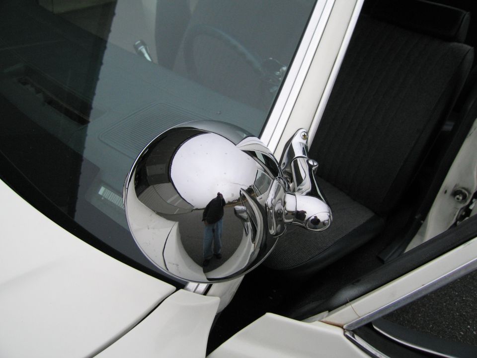 Reflection in a round chrome side mirror shows a person standing outside a white car