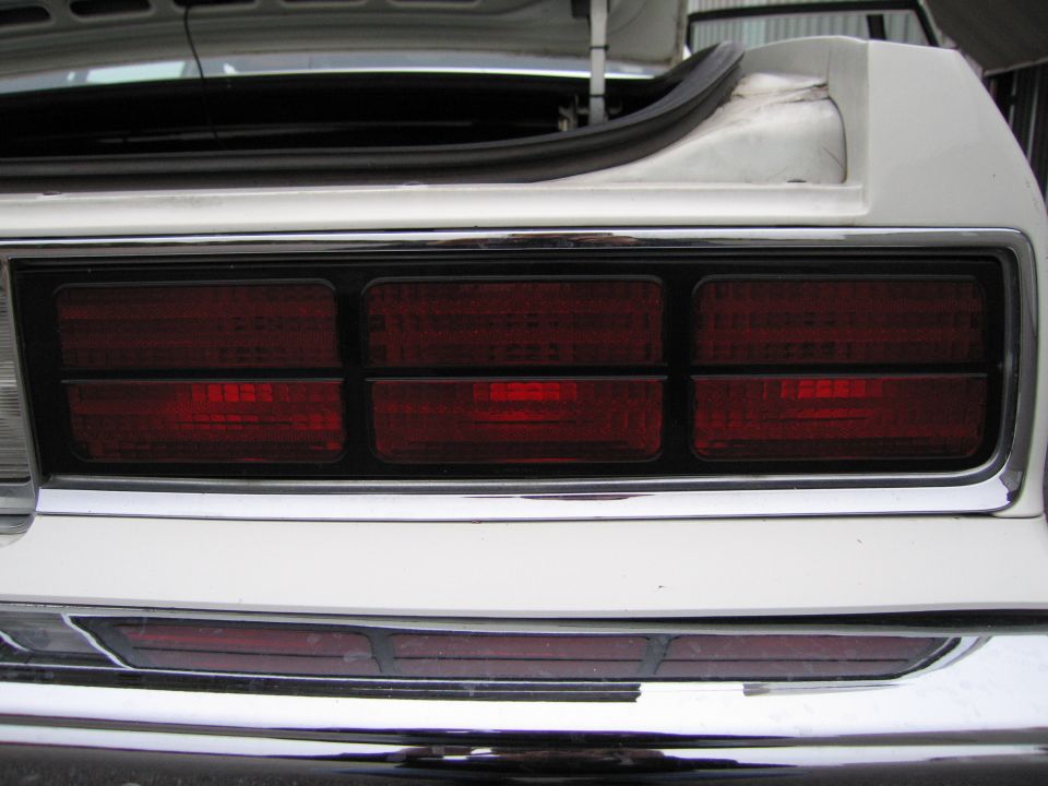 White car's rear end showing triple rectangular taillights on each side, chrome bumper, and open trunk