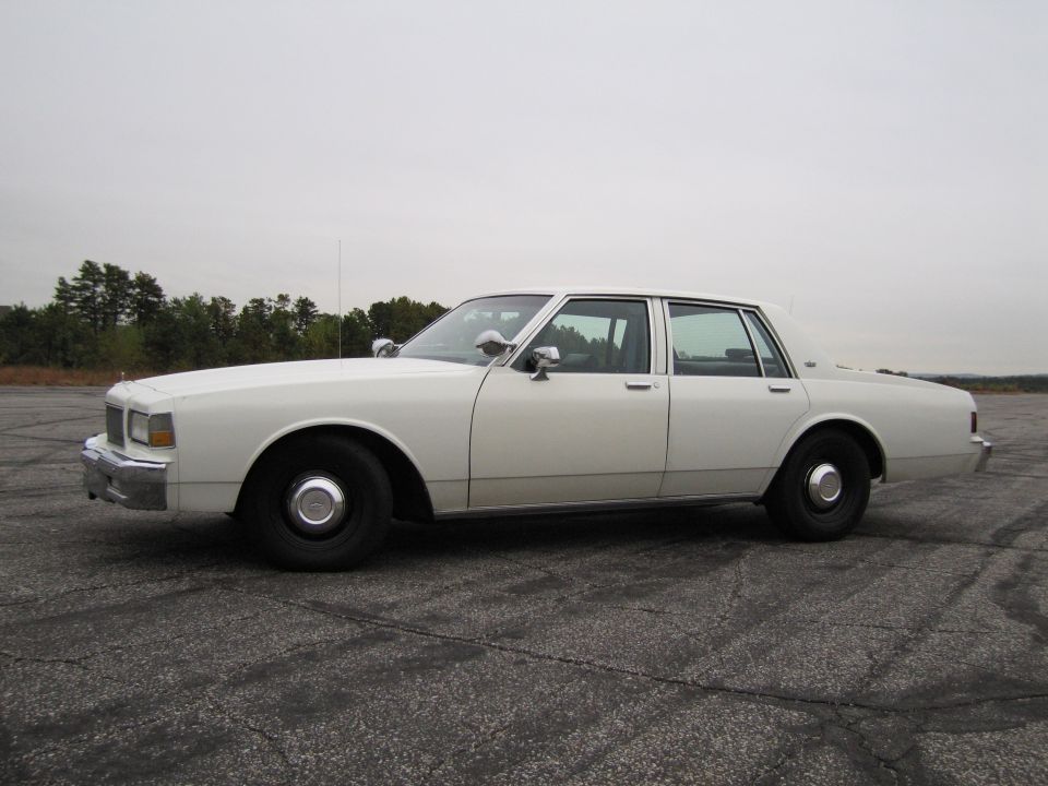 White sedan parked on asphalt, side view. The car has chrome mirrors and black wheels