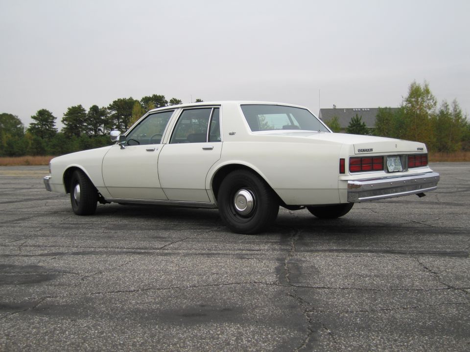 White sedan, likely 1980s Chevrolet Caprice, parked outdoors. Rear view shows taillights and chrome bumper