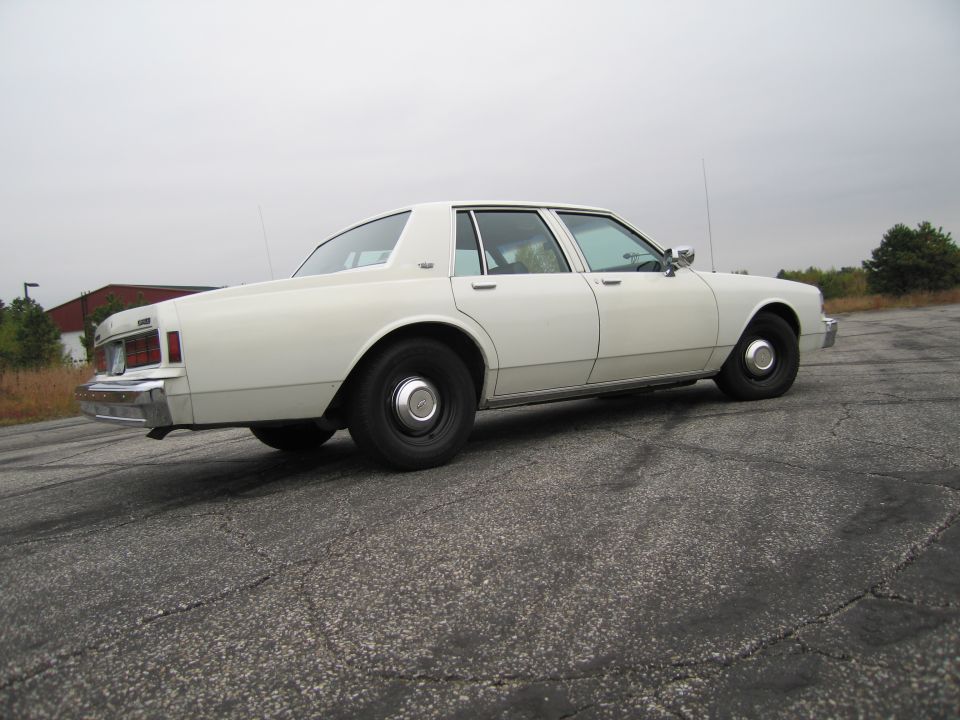 White sedan, likely 1980s Chevrolet Caprice, parked on cracked asphalt lot
