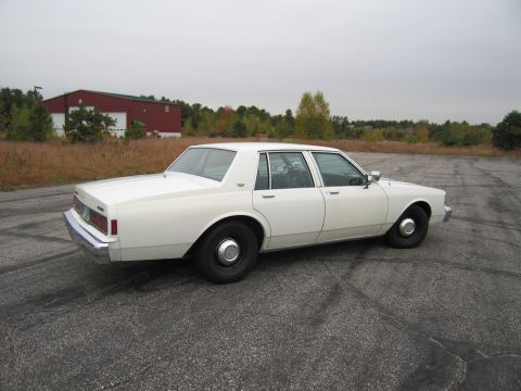 1991 Ford LTD Crown Victoria - This white sedan is parked outdoors on a cloudy day, with a red building and autumn foliage visible behind it.