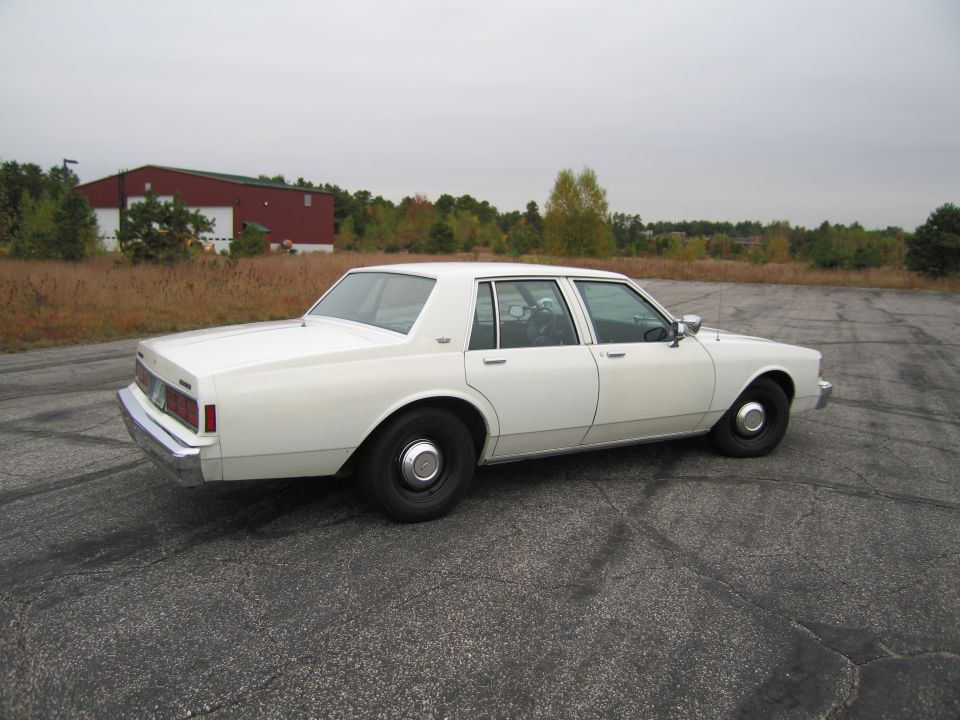 White sedan parked on cracked asphalt with a red building and trees in the background