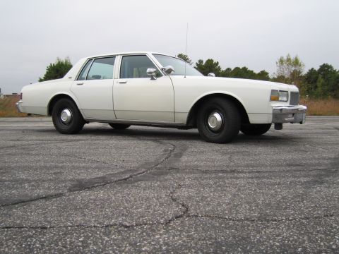 1991 Ford LTD Crown Victoria - Side profile of a white sedan, likely from the 1980s, parked on a cracked asphalt surface.