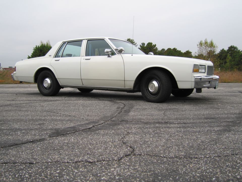 White sedan parked on asphalt, side view. Appears to be a police or taxi model from the 1980s