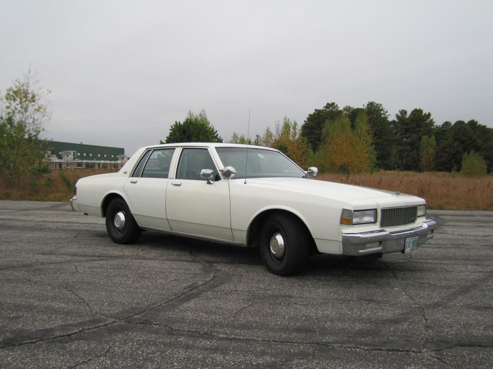 White sedan parked on cracked asphalt, front 3/4 view. Chrome grille and bumper visible