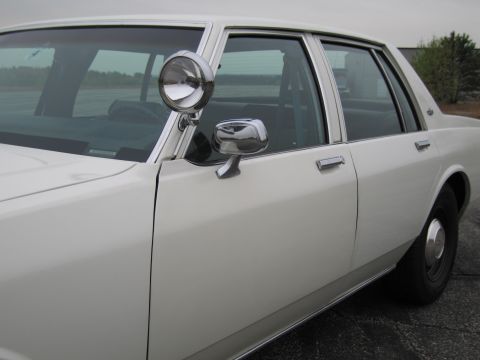 1991 Ford LTD Crown Victoria - This white sedan, likely a 1990 model, is viewed from the side, highlighting its police-style spotlight and chrome accents.