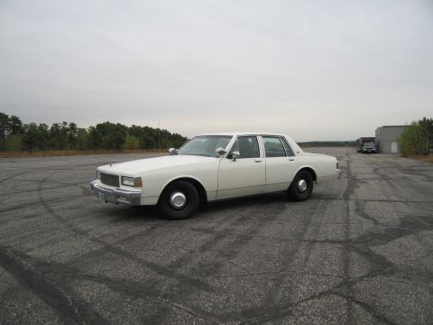1991 Ford LTD Crown Victoria - A white sedan sits on an empty asphalt lot under an overcast sky.