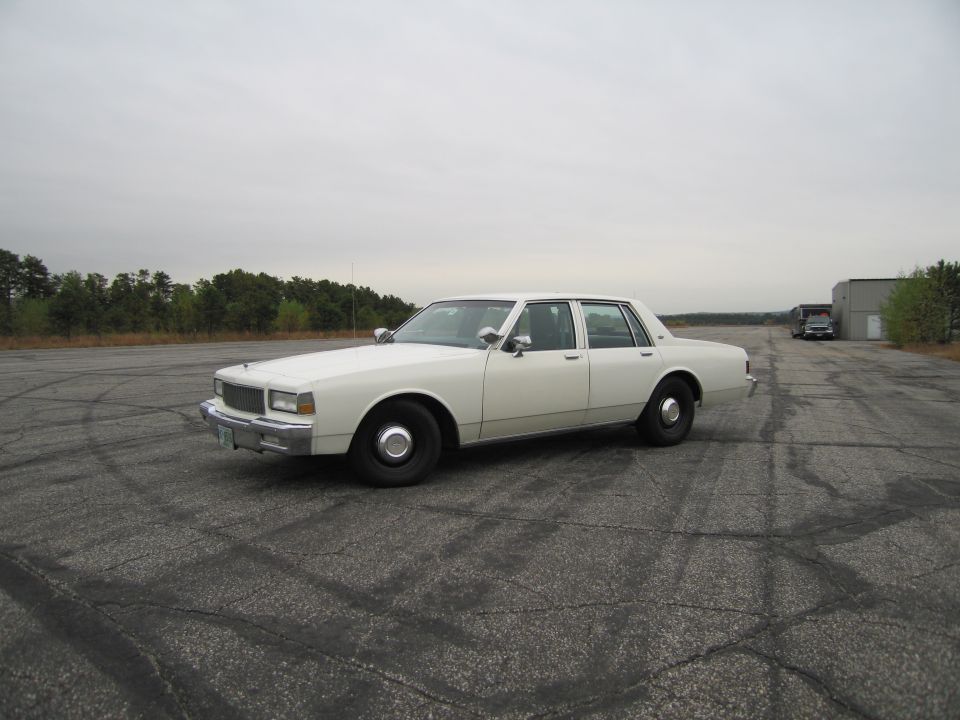 White sedan parked on cracked asphalt with trees in the background