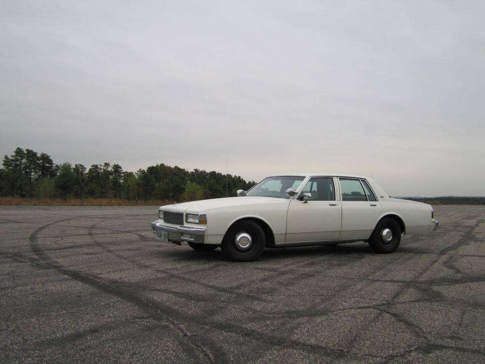 White 1980s sedan with black wheels parked on cracked asphalt with tire marks