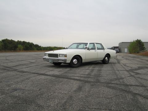1991 Ford LTD Crown Victoria - A white sedan sits on a desolate asphalt lot under an overcast sky.