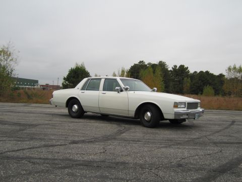 1991 Ford LTD Crown Victoria - A white 1980s Chevrolet Caprice sedan is parked on a cracked asphalt lot with trees in the background.
