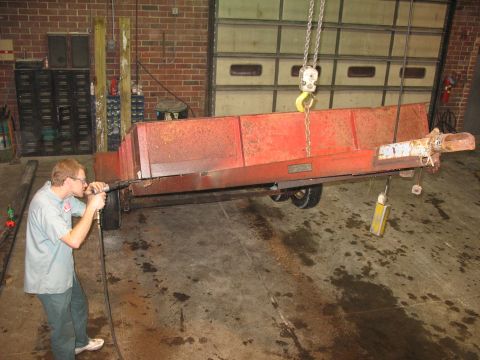 1900 Ford C-350 (Centurion Conversions) - A worker uses a tool on a red trailer that is being lifted by a crane inside a garage.