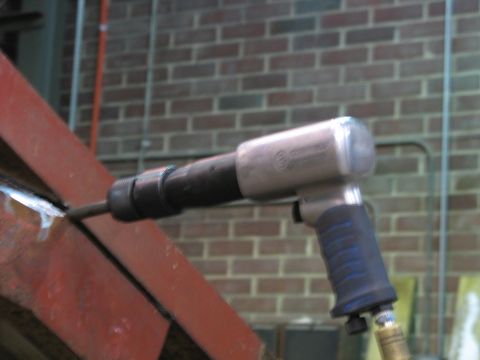 1900 Ford C-350 (Centurion Conversions) - A pneumatic chipping hammer is used on a red metal beam, with a brick wall in the background.