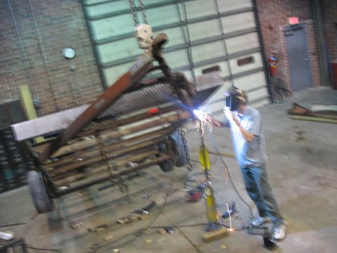 1900 Ford C-350 (Centurion Conversions) - A person welds a metal trailer in a workshop while it is suspended by a crane.