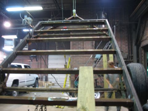 1900 Ford C-350 (Centurion Conversions) - A metal structure with stairs is suspended by a chain hoist in a workshop, with a white pickup truck visible in the background.