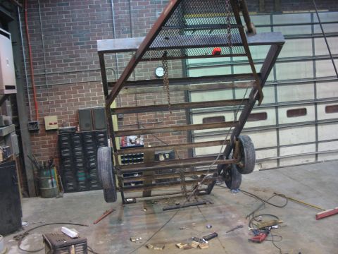 1900 Ford C-350 (Centurion Conversions) - A metal trailer frame is suspended by chains in a workshop, with tools and equipment scattered on the floor.