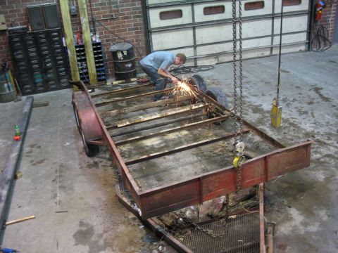 1900 Ford C-350 (Centurion Conversions) - A man grinds sparks off a trailer frame in a workshop. The trailer appears to be undergoing repairs or modifications.
