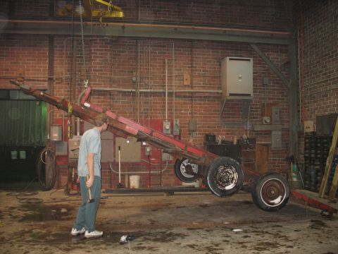 1900 Ford C-350 (Centurion Conversions) - A man inspects a red trailer frame suspended by a crane in a workshop setting.