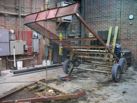 1900 Ford C-350 (Centurion Conversions) - A metal structure is suspended above a trailer chassis in a workshop setting.