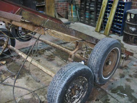 1900 Ford C-350 (Centurion Conversions) - A rusty trailer chassis with two wheels is visible in a workshop setting, with various tools and barrels in the background.