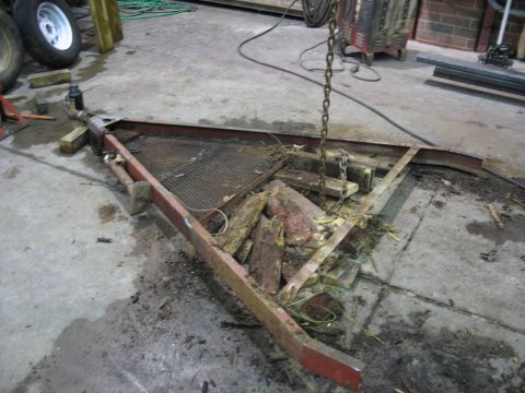 1900 Ford C-350 (Centurion Conversions) - A damaged red trailer frame with a mesh deck and debris is suspended by a chain in a workshop.
