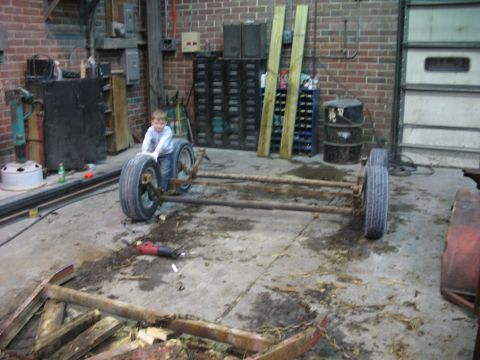 1900 Ford C-350 (Centurion Conversions) - A young boy pushes a bare vehicle frame with wheels in a cluttered garage setting.