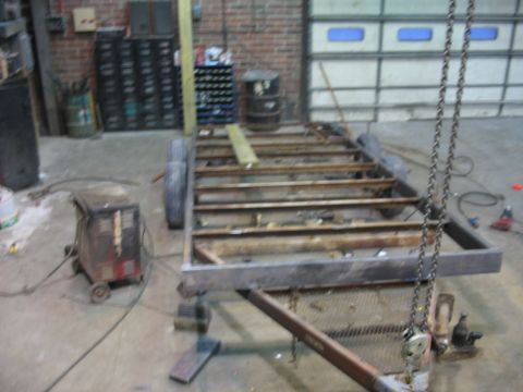 1900 Ford C-350 (Centurion Conversions) - A bare trailer frame sits in a workshop, with a welding machine nearby and a large garage door in the background.