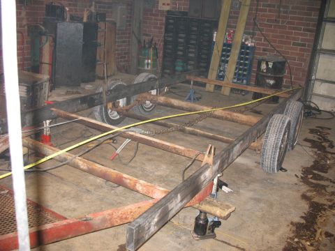 1900 Ford C-350 (Centurion Conversions) - A rusty trailer frame is shown in a garage, supported by hydraulic jacks and dual axles with tires.
